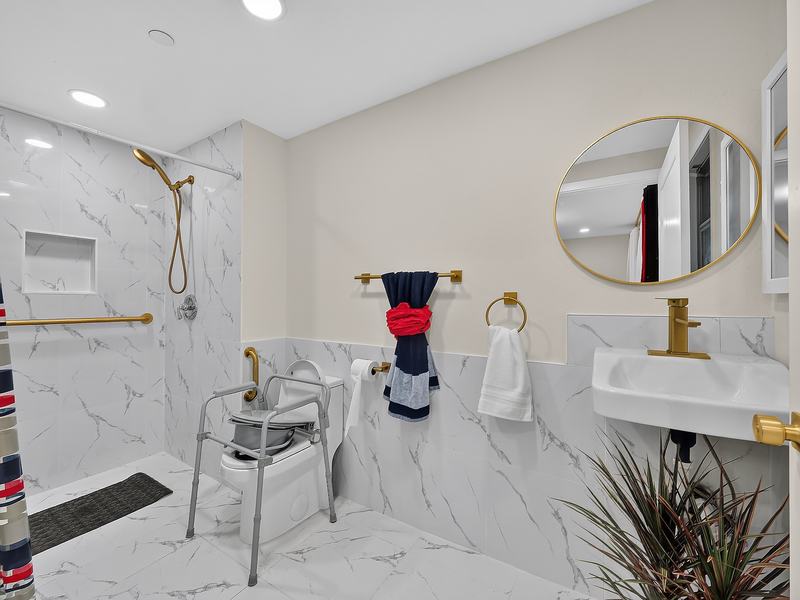 A marble-tiled walk-in shower with a handheld sprayer, next to a raised toilet seat with rails