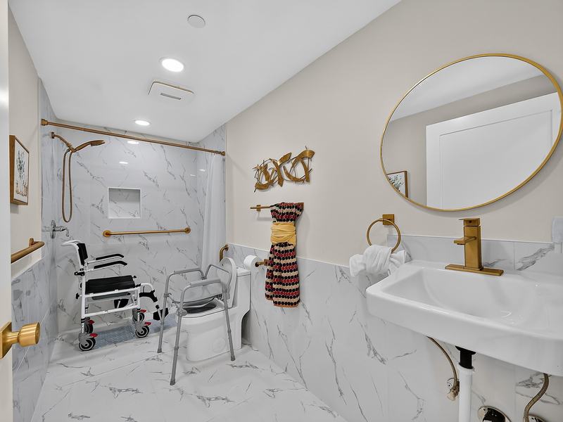 A bathroom with a roll-in shower, shower wheelchair, grab bars, and bedside commode